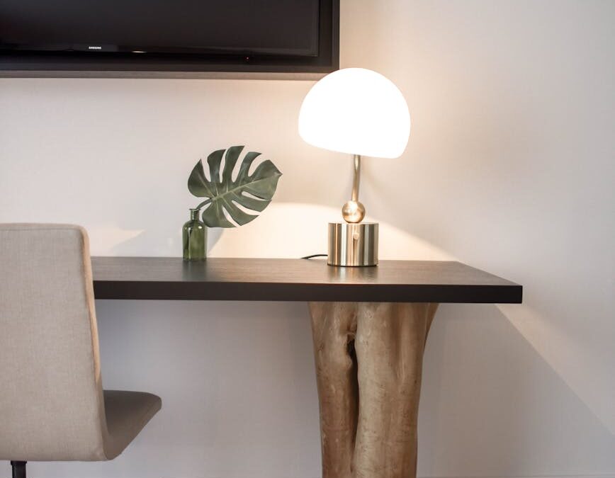 stainless steel base white shade table lamp on brown wooden desk near white painted wall with wall mounted flat screen t v