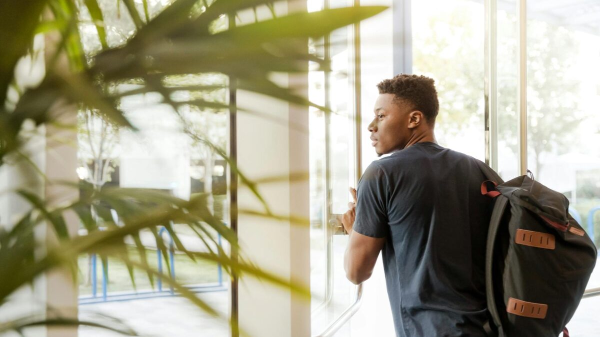 man looking outside window carrying black and brown backpack while holding his hand on window