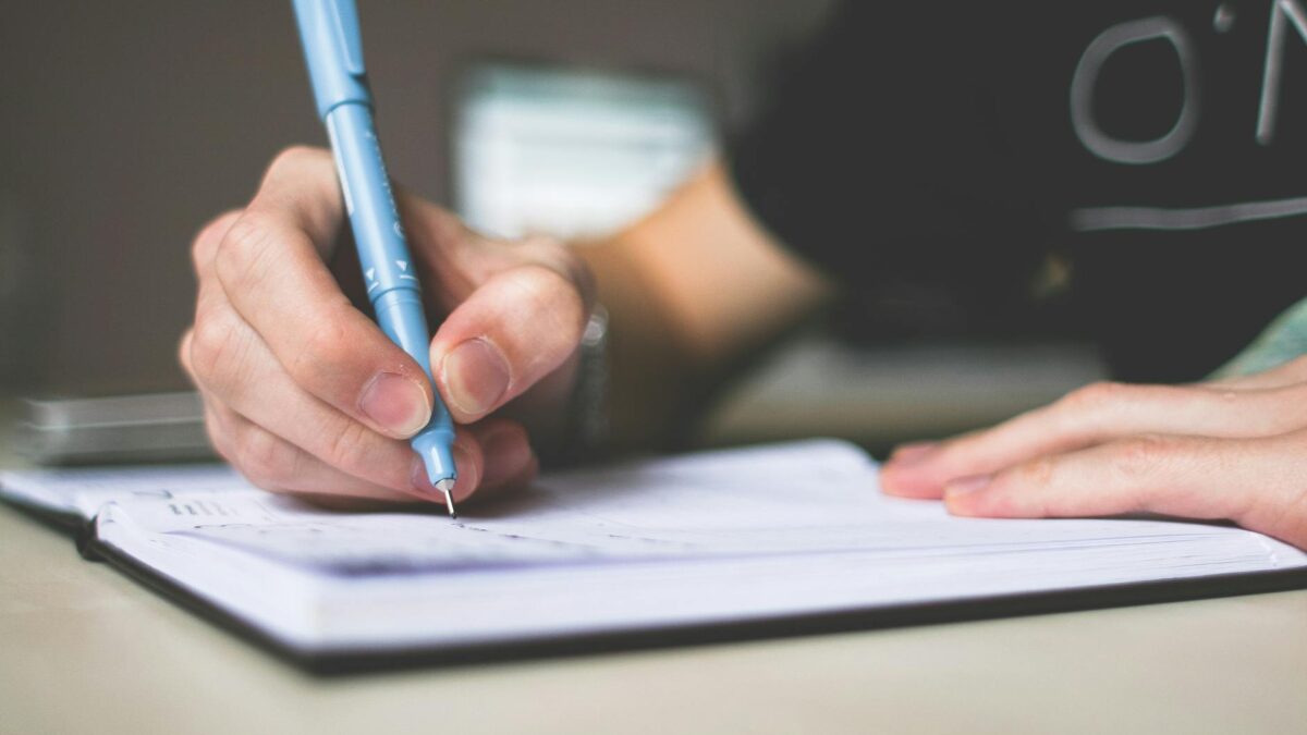 person holding blue ballpoint pen writing in notebook